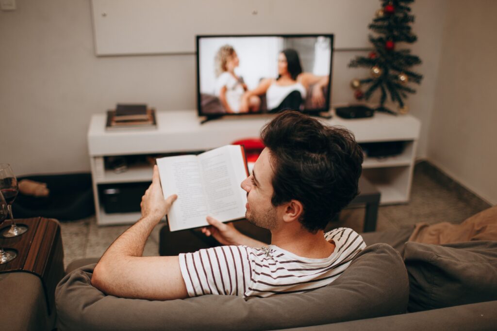 A man sits on a sofa reading a book in a cozy living room with a TV on.